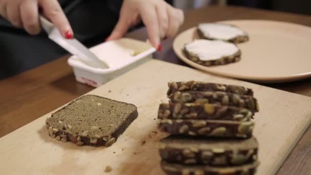 Een vrouwelijke hand spreidt donker roggebrood uit met roomkaas met een mes — Stockvideo