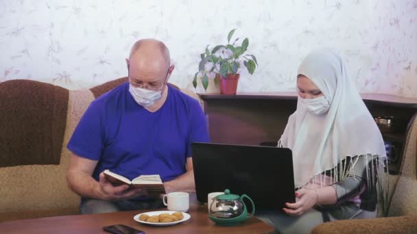 Muslimský pár v ochranných maskách karanténní pro prevenci koronaviru doma na gauči manžel čte knihu manželka pracuje na počítači. — Stock video