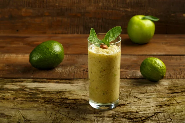 Un bicchiere di frullato verde con menta su un tavolo di legno su un tovagliolo vicino a frutta e noci . — Foto Stock
