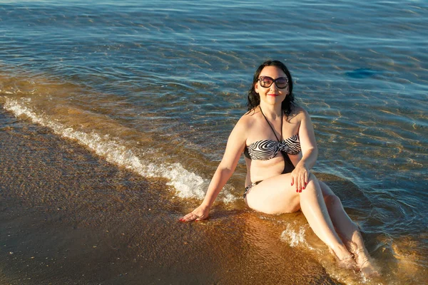 En brunett kvinna i baddräkt och glasögon sitter på stranden. — Stockfoto