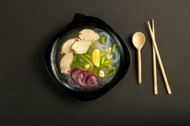 Siyah arka planda tavuklu bir tabak Vietnamlı Pho çorbası, kaşık ve çubuklarla..