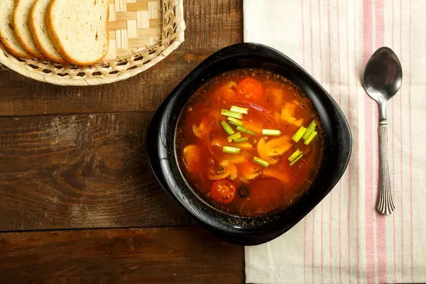 El plato negro de la sopa italiana de tomate con las patatas y las setas sobre la mesa de madera sobre la servilleta y la cuchara y el pan . — Foto de Stock