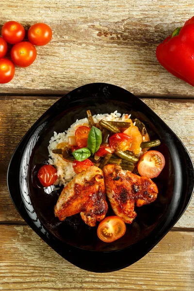用番茄酱烤鸡翅，在靠近蔬菜的木制桌子上的黑色盘子里放上米饭和蔬菜. — 图库照片