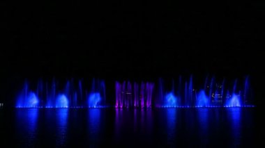 Çok renkli fıskiyelerin ışıl ışıl fıskiyeleri gece gökyüzünün arka planında suya yansıyor.