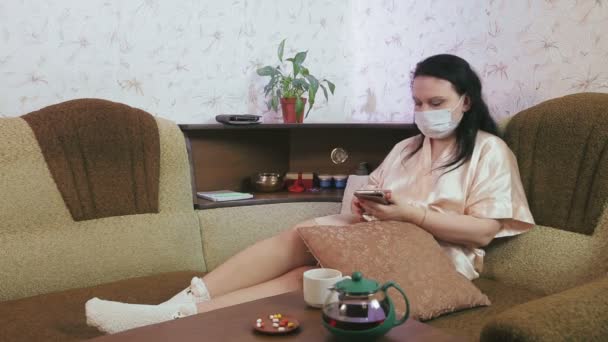 A young woman in a home dressing gown and a medical mask at home in quarantine self-isolation on the couch communicates in a messenger in a smartphone — Stock Video