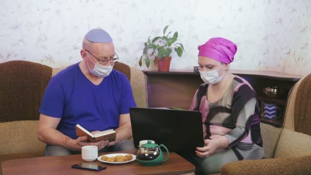 Ebrei coppia sposata in maschere protettive in quarantena per la prevenzione del coronavirus a casa sul divano marito legge un libro moglie lavora al computer . — Video Stock