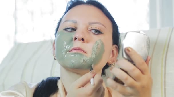 Una mujer en casa se pone una máscara de arcilla azul en la cara. . — Vídeo de stock