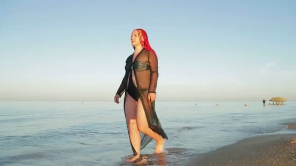 Žena v plavkách a černý župan na moři kráčí podél pobřeží — Stock video