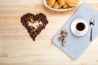 Tahta bir masanın üzerindeki peçetede bir fincan kahve ve kalp şeklinde dağınık bir kahve sarılığı. Yatay fotoğraf