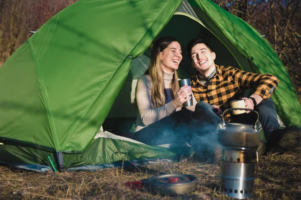 Feliz joven pareja enamorada de la naturaleza sonriendo, tomando té y café en la tienda de campaña cerca de un fuego. actividades al aire libre para familias jóvenes — Foto de Stock