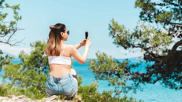 young woman traveler in sunglasses taking photo of sea scenery in mid day. blue sea and pine trees in croatia