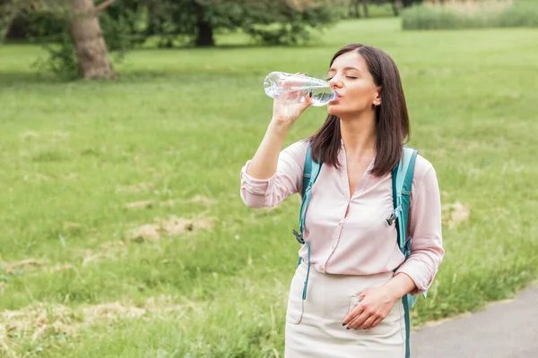 Весной молодая женщина в повседневной одежде пьет воду из пластиковой бутылки в парке — стоковое фото