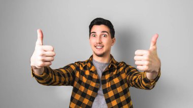 Sarı gömlekli yakışıklı genç adam iki eliyle baş parmağını kaldırıyor. Gri arka plana karşı jest gibi.