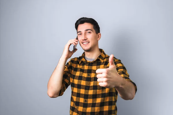 Handsome young man or student in yellow shirt with smile on face gets good news by phone and shows like sign, big thumb up — Stock Photo, Image