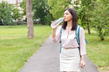 Vücut sıvısı. Kadın, günlük giysiler ve sırt çantasıyla parkta bir şişeden su içiyor.