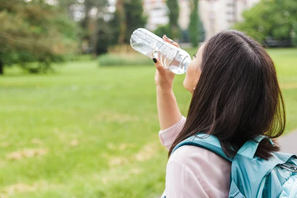 Közelkép nő alkalmi ruhákban és hátizsák ivóvíz egy palackból a parkban. testhidratáció — Stock Fotó