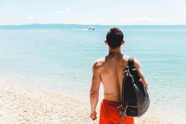 Sırt çantasıyla tek başına okyanusa açılan bir tekneye bakan genç bir gezgin..