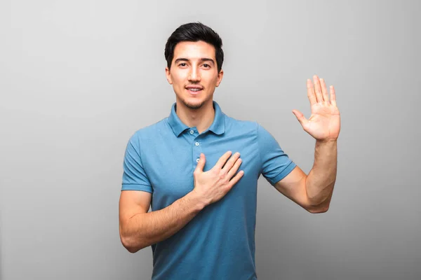 I swear. handsome young man in blue shirt with hand on chest giving oath against grey background. studio shoot — Stock Photo, Image