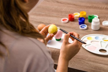 Paskalya havası. Kapatın genç kadın elleri paskalya yumurtalarını fırçayla boyuyor ve evde ahşap masanın üzerine pastel renkte balıklar çiziyor..