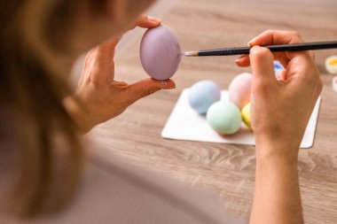 Paskalya havası. Kapatın genç kadın elleri paskalya yumurtalarını fırçayla boyuyor ve evde ahşap masanın üzerine pastel renkte balıklar çiziyor..