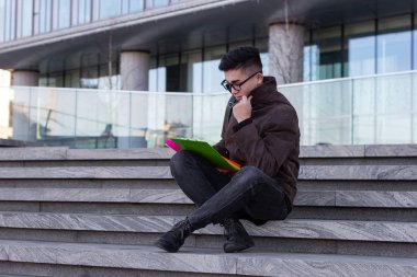 Üniversitenin basamaklarında oturan genç Asyalı erkek öğrenci kitap okuyup şöyle düşündü: