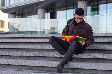 Genç Asyalı erkek öğrenci üniversitenin merdivenlerinde oturup kitap okuyor.