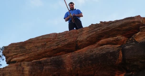 の崖の上に立っている間の腰に自分のロープを調整する運動選手の低い角度表示 — ストック動画