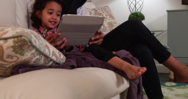 若い混合レースの母親のフロントビューでは 彼女の若い娘と一緒に座ってソファに座って タブレットコンピュータを使用してヒジャーブを身に着けています — ストック動画
