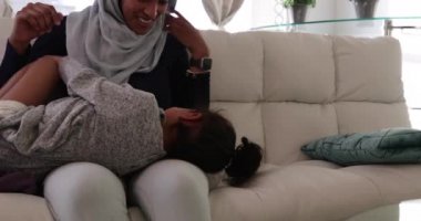 Genç bir melez annenin, genç kızıyla oturma odasında tesettürde otururken, birlikte gülümseyip oynaşırken, annesinin kucağında yatan kızın ön görüntüsü.