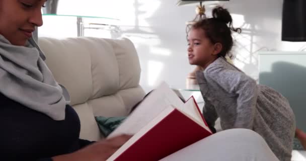 サイドビューアップアップの若いです混合レースの母親身に着けていますHijabとともに彼女の若い娘でシッティングルーム ソファに座って本を読んで — ストック動画