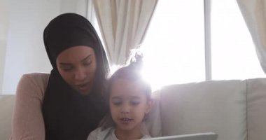 Genç melez bir kadının, genç kızıyla oturma odasında tesettüre oturup tablet bilgisayar kullanması ön planda.