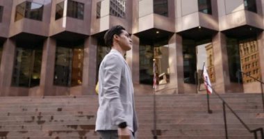 Tablet bilgisayar taşıyan, zamanı kontrol eden ve arka planda binalarla şehir caddesinde yürüyen melez bir adamın yan görüntüsü.