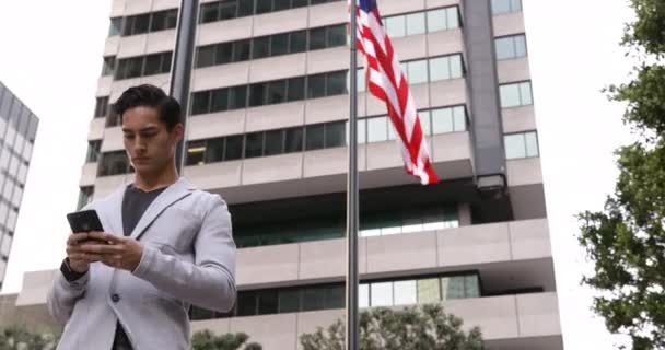 一个年轻的混血种族男子用智能手机站在城市街道上 以建筑物为背景的低视角前看 — 图库视频影像