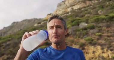 Sahilde kırsalda koşan ve su şişesinden su içen beyaz bir adamın ön görüntüsü.