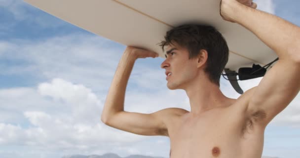 一个赤身裸体的白人年轻人站在海滩上 头顶冲浪板 凝视着大海 身后是蓝天 动作缓慢 — 图库视频影像