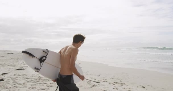 ウェットスーツを着てサーフボードを持って海に向かって走るビーチで若い白人白人男性のサイドビュー スローモーション — ストック動画