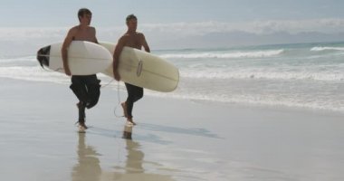 Beyaz gömleksiz bir baba ve genç yetişkin oğlunun dalgıç kıyafeti giyip, sahilde sörf tahtası taşıması, ağır çekim...
