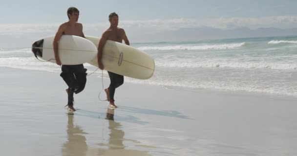 一个赤身裸体的高加索父亲和他年轻的成年儿子穿着湿衣 背着冲浪板在阳光下沿着海滩奔跑 动作缓慢 — 图库视频影像