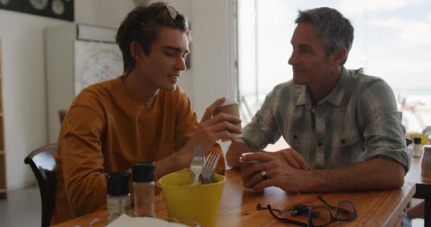 カフェでコーヒーを飲みながらゆっくりと話をするテーブルに座っている白人の父と彼の若い大人の息子のフロントビュー — ストック動画