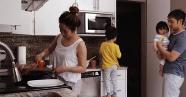 Çinli Asyalı bir ailenin yan görüntüsü, mutfakta birlikte meşgul olan anne tavayla yemek pişiriyor, baba genç kızını kollarına alıyor ve mikrodalga fırını kullanmak için taburede duran oğluna yardım ediyor.