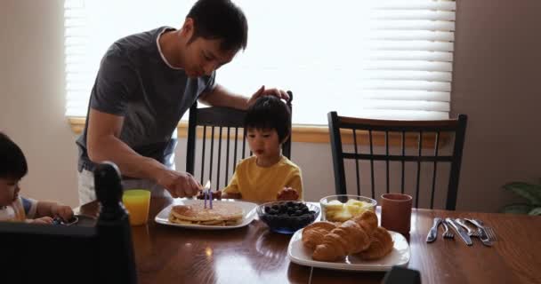 一个亚洲中国家庭在他们的餐厅里 父母站在他们年幼的儿子和女儿坐的桌子旁边 父亲在生日蛋糕上点蜡烛为他的儿子 — 图库视频影像