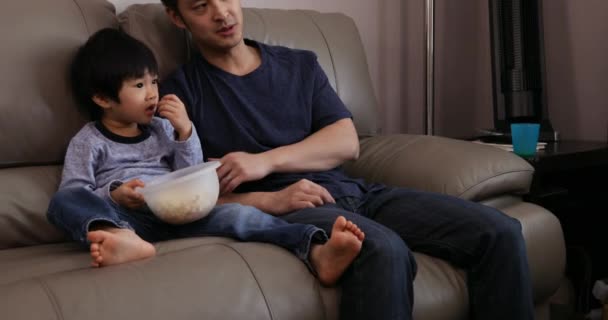 彼らのリビングルームで中国のアジア人男性と彼の若い息子のフロントビューを閉じると 一緒にソファの上にテレビを見て座って 息子は彼が保持しているボウルからポップコーンを食べて — ストック動画