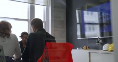 Modern bir ofiste, melez bir iş kadınıyla yapılan toplantı sırasında masa başında oturan beyaz bir erkek ve kadının arka planda, adamın koltuğunun arkasında bir yeleği var. Yansımaları olan camdan bir duvarın içinden.