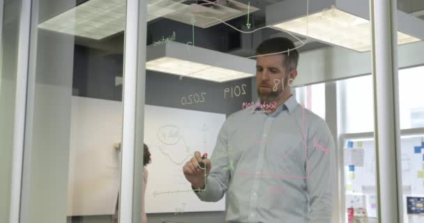 現代的なオフィスで働く白人男性のビジネスクリエイティブのフロントビュー ガラスの壁に色のペンでグラフや図を描く 2人の女性の同僚がバックグラウンドで作業を表示されます ガラスの壁を通して見ると — ストック動画