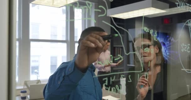 Vue de face d'un homme d'affaires afro-américain travaillant avec une femme d'affaires caucasienne dans un bureau moderne, debout et ayant une discussion, dessinant des graphiques et des diagrammes sur un mur de verre ensemble, un collègue masculin travaillant en arrière-plan 