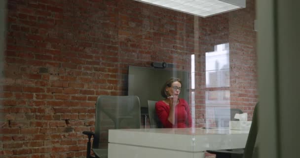 現代的なオフィスでスマートフォンで話している机に座ってタブレットPcを見ている白人女性ビジネスの専門家の側のビュー 反射とガラスの壁を通して見られます — ストック動画