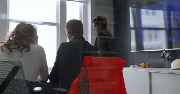 会議室の机に腰を下ろしていた白人男性と女性の後ろ姿と 現代のオフィスで人種の混じった実業家の女性が加わり 椅子の後ろに腰を下ろしている 反射のあるガラスの壁を通して見る — ストック動画