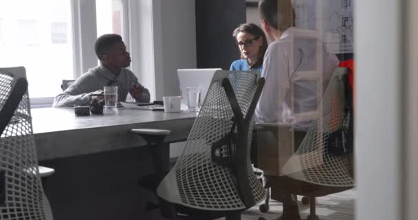 ラップトップを使用して白人女性の側のビュー 現代のオフィスでの会議中にテーブルに座って混合人種の男とアフリカ系アメリカ人の男 一緒に壁に取り付けられた画面を話し 反射のあるガラスの壁を通して見る — ストック動画