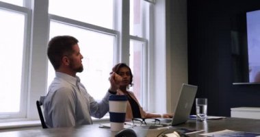 Modern bir ofiste çalışan bir grup iş arkadaşının yan görüntüsü. Toplantı odasında oturmuş duvarda asılı duran melez bir kadın meslektaşla görüntülü bir görüşme yapıyorlar.