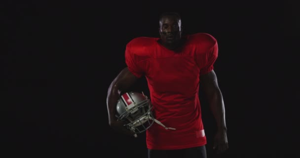 彼の腕の下にヘルメットで立って チームの制服 パッドと目の黒を身に着けているアフリカ系アメリカ人男性アメリカンフットボール選手のフロントビュー — ストック動画
